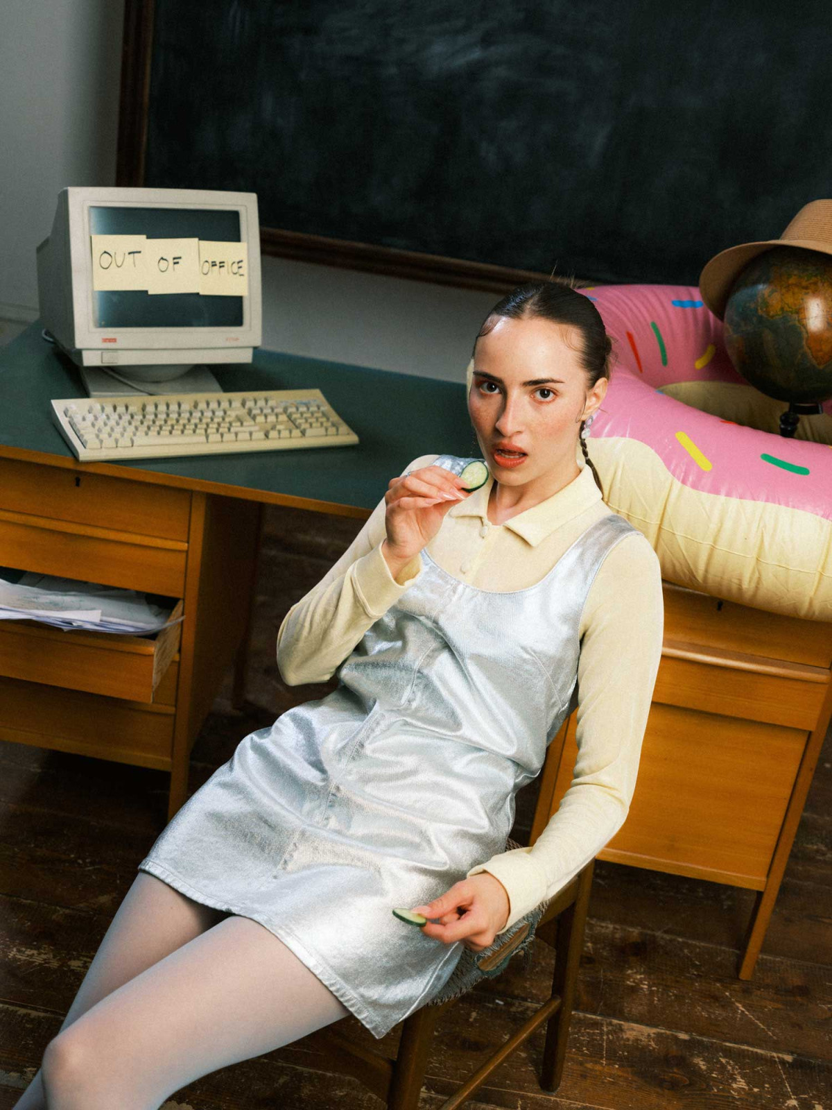 Woman in bright clothing leaning against chair holding cucumber slice in hand in classroom environment