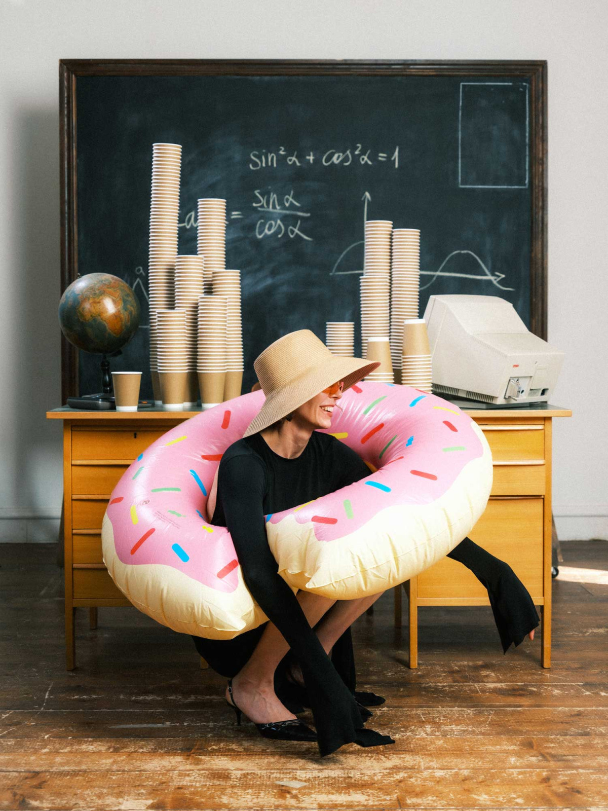 Woman in classroom environment squatting with doughnut shaped floatie in front of a vintage table covered with towers of coffee cups on chalkboard backdrop