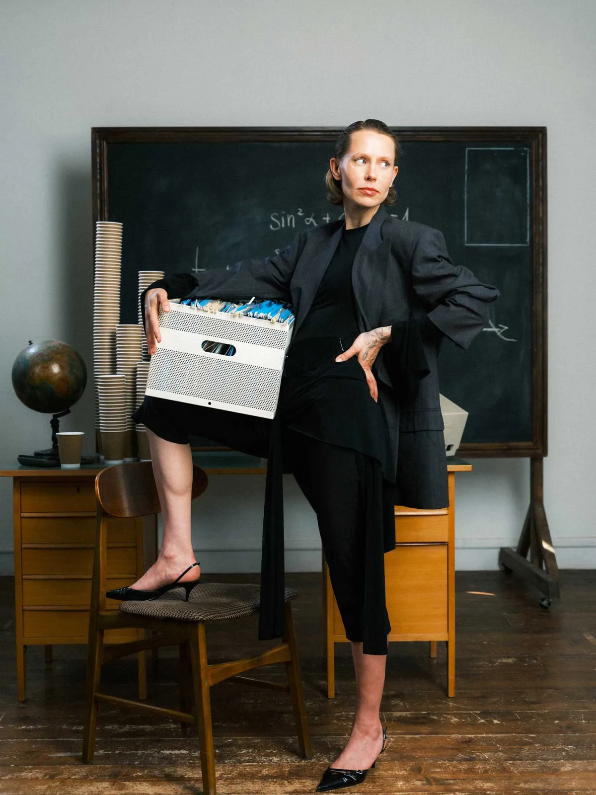 Woman in dark clothing resting one leg on top of a chair holding box with files in front of a vintage table covered with towers of coffee cups on chalkboard backdrop in classroom environment