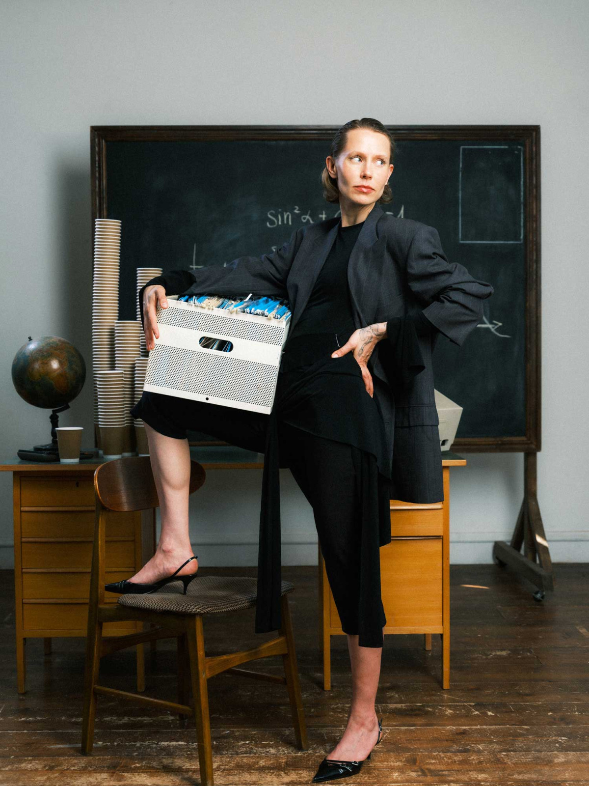 Woman in dark clothing resting one leg on top of a chair holding box with files in front of a vintage table covered with towers of coffee cups on chalkboard backdrop in classroom environment