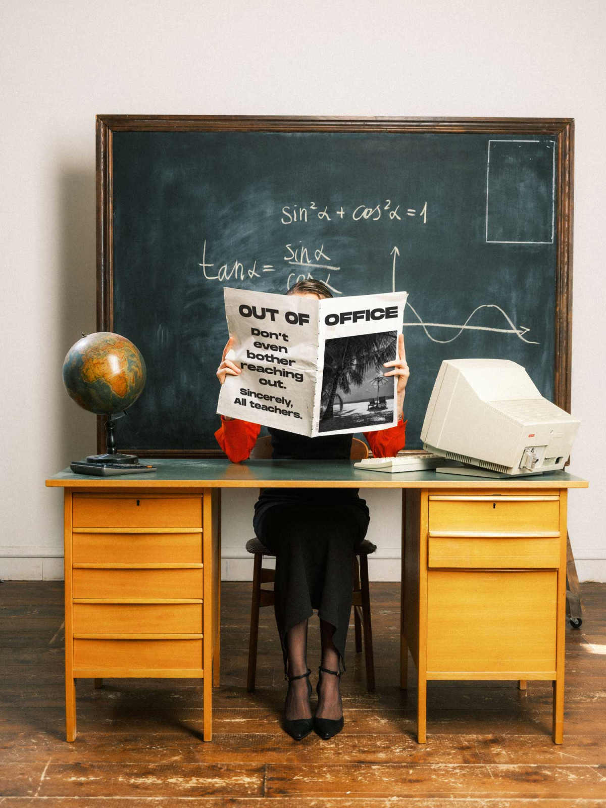 Woman sitting behind vintage table in classroom environment holding up a newspaper with text saying Out Of Office